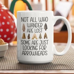 May include: White ceramic mug with a black inscription that reads "Not all who wander are lost. Some are just looking for arrowheads." The mug has a black handle and is decorated with images of arrowheads.