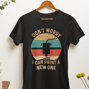 May include: Black t-shirt with a vintage-style graphic of a 3D printer. The graphic has a red, orange, and yellow sunset background with the text "Don't Worry I Can Print A New One".