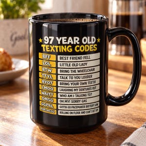 Może przedstawiać: Czarny ceramiczny kubek z żółto-białym napisem 97 YEAR OLD TEXTING CODES, po którym następuje lista humorystycznych akronimów i ich znaczeń, w tym BFF dla Best Friend Fell, LOL dla Little Old Lady oraz ROFLACGU dla Rolling On Floor And Can't Get Up.