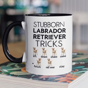 Könnte beinhalten: Eine weiße Kaffeetasse mit schwarzem Henkel und schwarzem Rand. Die Tasse trägt den Text "Stubborn Labrador Retriever Tricks" in schwarzer Schrift. Unter dem Text befinden sich sechs Bilder von Labrador Retrievern mit dem Text "sit", "down", "shake", "come", "fetch", "roll over" und "stay" unter jedem Bild.