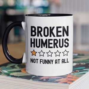 May include: White coffee mug with a black handle and black rim. The mug has the text "BROKEN HUMERUS" in black and a one-star rating with the text "NOT FUNNY AT ALL" in black.