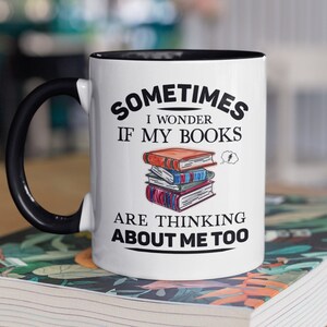 Pode incluir: Caneca de cerâmica branca com alça e borda pretas. A caneca tem um gráfico preto que diz "Às vezes me pergunto se meus livros estão pensando em mim também." O gráfico também inclui uma pilha de livros com um balão de pensamento sobre eles.