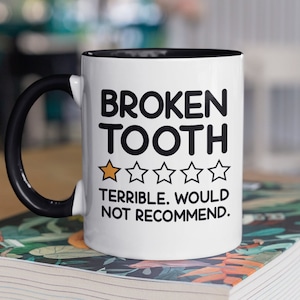 May include: White ceramic mug with a black handle and black rim. The mug has a black graphic that says "BROKEN TOOTH" with a one-star rating and the text "TERRIBLE. WOULD NOT RECOMMEND."