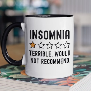 May include: White coffee mug with a black handle and black rim. The mug has black text that reads "INSOMNIA" with a one-star rating and the text "TERRIBLE. WOULD NOT RECOMMEND."