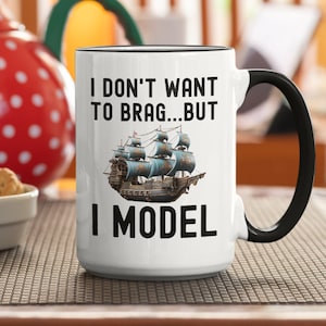 May include: White ceramic coffee mug with a black handle. The mug has black text that reads "I don't want to brag...but I model" and a picture of a ship.
