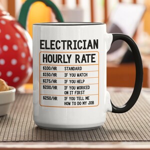 Peut inclure: Une tasse à café en céramique blanche avec une poignée noire. La tasse a un bord noir et un texte noir qui dit "ELECTRICIAN HOURLY RATE" avec une liste de tarifs horaires et les raisons de ces tarifs.
