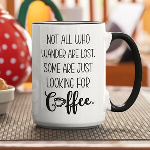 May include: White ceramic coffee mug with a black handle. The mug has black text that reads "Not all who wander are lost. Some are just looking for coffee." with a small coffee cup icon.
