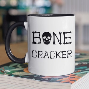 May include: White ceramic mug with a black handle and black rim. The mug has a black skull and crossbones design with the text "BONE CRACKER" printed on it.