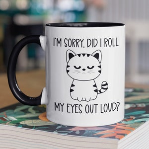 May include: White ceramic coffee mug with a black handle and black rim. The mug has a black and white cartoon cat with the text "I'm sorry, did I roll my eyes out loud?"