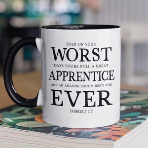Puede incluir: Taza de cerámica blanca con interior y asa negros. La taza presenta el texto "EVEN ON YOUR WORST DAYS YOU'RE STILL A GREAT APPRENTICE AND AN AMAZING PERSON. DON'T YOU EVER FORGET IT!" en letras negras. La taza está sobre un libro.