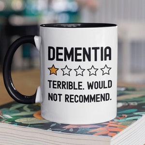 Puede incluir: Taza de café blanca con asa negra y borde negro. La taza tiene texto negro que dice "Dementia" con una calificación de una estrella y el texto "Terrible. No lo recomendaría."