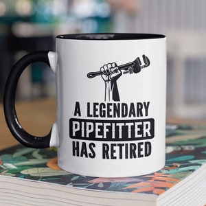 May include: A white coffee mug with a black handle and black rim. The mug features a black and white illustration of a hand holding a wrench. The text "A Legendary Pipefitter Has Retired" is printed in black on the mug.