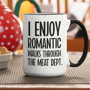 May include: White ceramic mug with black handle and rim. The mug features the text "I ENJOY ROMANTIC WALKS THROUGH THE MEAT DEPT." in black, bold font. A red and white polka-dot teapot and a small bowl of cookies are in the background.
