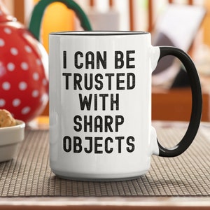 May include: White ceramic coffee mug with a black handle. The mug has black text that reads "I can be trusted with sharp objects".