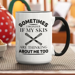 Op de afbeelding: Een witte keramische mok met een zwarte handgreep. De mok heeft zwarte tekst die luidt "Sometimes I wonder if my skis are thinking about me too." De tekst is omgeven door een zwarte rand. Een paar zwarte ski's zijn gekruist in het midden van de mok.