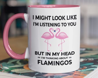 May include: White ceramic mug with a pink rim and handle. The mug has a black graphic with two pink flamingos and the text "I might look like I'm listening to you, but in my head I'm thinking about flamingos."