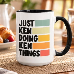 Könnte beinhalten: Ein weißer Keramikbecher mit schwarzem Henkel und schwarzem Innenrand. Auf der Vorderseite steht der schwarze Text "JUST KEN DOING KEN THINGS" vertikal untereinander, wobei jede Zeile von einem horizontalen rechteckigen Farbblock in Salbei, Gelb, Gold, Orange und Rot begleitet wird.