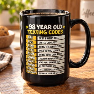 Può includere: Una tazza in ceramica nera con il testo giallo 98 YEAR OLD TEXTING CODES seguito da un elenco di acronimi umoristici e i loro significati, tra cui BFF per BEST FRIEND FELL, LOL per LITTLE OLD LADY e BYOT per BRING YOUR OWN TEETH.