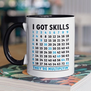 May include: White ceramic coffee mug with a black rim and handle. The mug has black text that reads "I GOT SKILLS" and a grid of numbers that multiply by the row number. The text at the bottom reads "THEY'RE MULTIPLYIN".