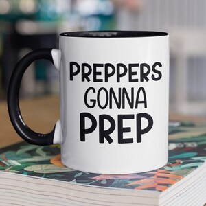 May include: White coffee mug with a black handle and black rim. The mug has the text "Preppers Gonna Prep" in black lettering.