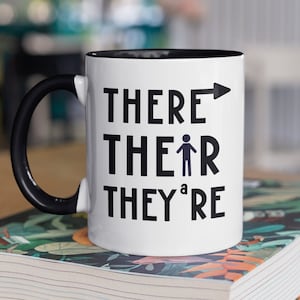 Puede incluir: Una taza de café blanca con asa negra y borde negro. La taza tiene las palabras "THERE", "THEIR" y "THEY'RE" impresas en negro. La palabra "THEIR" tiene una figura de palo a su lado. La taza está encima de un libro.