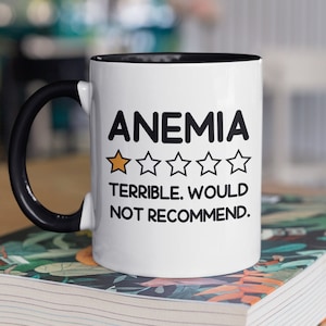 Può includere: Una tazza da caffè bianca con un manico nero e un bordo nero. La tazza ha una grafica in bianco e nero che dice "Anemia" con una valutazione a una stella e il testo "Terribile. Non lo consiglio."