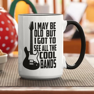 May include: White ceramic coffee mug with a black handle. The mug has a black and white graphic of an electric guitar and the text "I may be old but I got to see all the cool bands".