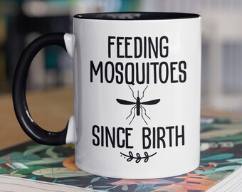 May include: White coffee mug with a black handle and black rim. The mug has black text that reads "Feeding Mosquitoes Since Birth" with a mosquito graphic.