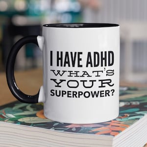 Op de afbeelding: Een witte koffiemok met een zwarte handgreep en een zwarte rand. De mok heeft zwarte tekst die luidt "Ik heb ADHD Wat is jouw superpower?" De mok staat op een boek.