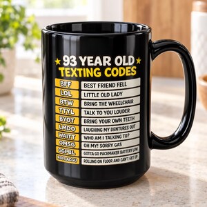 Könnte beinhalten: Ein schwarzer Keramikbecher mit dem gelb-weißen Text 93 YEAR OLD TEXTING CODES, gefolgt von einer Liste humorvoller Akronyme und deren Bedeutungen, darunter BFF Best Friend Fell, LOL Little Old Lady und ROFLACGU Rolling On Floor And Can't Get Up.