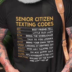 May include: Black t-shirt with a white graphic print of a list of senior citizen texting codes. The text includes abbreviations like BFF, LOL, BTW, TTYL, BYOT, LMDO, WAITT, OMSG, GGPBL, and ROFLACGU. Each abbreviation is followed by a description of what it means.