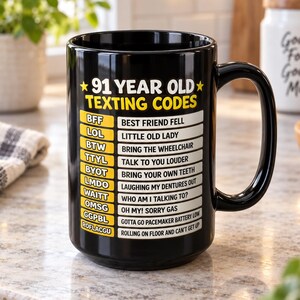 Puede incluir: Una taza de cerámica negra con el texto amarillo 91 YEAR OLD TEXTING CODES y una lista de acrónimos humorísticos y sus significados, incluyendo BFF para Best Friend Fell, LOL para Little Old Lady y ROFLACGU para Rolling On Floor And Can't Get Up.
