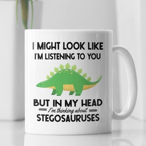 Può includere: Una tazza da caffè in ceramica bianca con un dinosauro stegosauro verde a cartoni animati e il testo "Potrei sembrare che ti stia ascoltando, ma nella mia testa sto pensando agli stegosauri."