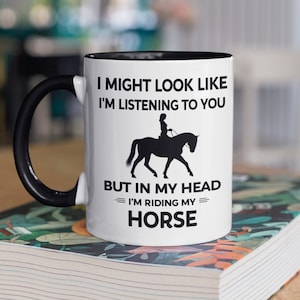 May include: White ceramic coffee mug with a black handle and black rim. The mug has a black silhouette of a person riding a horse and the text "I might look like I'm listening to you but in my head I'm riding my horse."