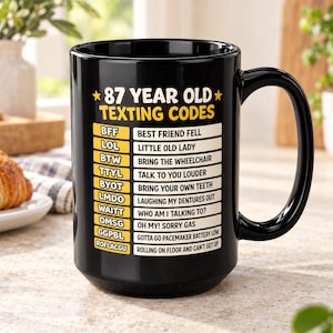 May include: A black ceramic mug featuring the yellow and white text 87 YEAR OLD TEXTING CODES followed by a list of humorous acronyms and their meanings, such as BFF for BEST FRIEND FELL, LOL for LITTLE OLD LADY, and TTYL for TALK TO YOU LOUDER.