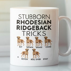 May include: White ceramic mug featuring a brown Rhodesian Ridgeback dog illustration and the text "Stubborn Rhodesian Ridgeback Tricks" with a list of dog tricks below the illustration: Sit, Down, Shake, Come, Fetch, Roll Over, Stay.
