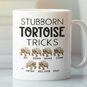 May include: White ceramic coffee mug with black text that reads "Stubborn Tortoise Tricks". The mug features a line of six tortoises with the words "Sit", "Down", "Shake", "Come", "Fetch", "Roll Over", and "Stay" written below them.