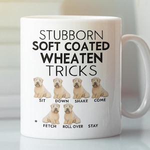 Może przedstawiać: Biały kubek ceramiczny z czarnym nadrukiem przedstawiającym psa rasy Soft Coated Wheaten Terrier. Tekst głosi "Stubborn Soft Coated Wheaten Tricks" i wymienia popularne komendy dla psów: Siad, Leżeć, Łapka, Chodź, Aport, Turlaj się, Zostań.
