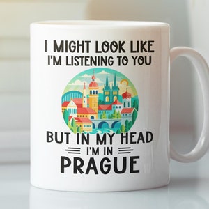 Op de afbeelding: Witte keramische koffiemok met een zwarte afbeelding van een stadsbeeld en de tekst "I might look like I'm listening to you but in my head I'm in Prague".