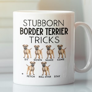 May include: White ceramic mug with a black and white design. The mug features a Border Terrier dog and the text "Stubborn Border Terrier Tricks". The text "Sit, Down, Shake, Come, Fetch, Roll Over, Stay" is listed below the dog illustrations.