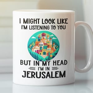 Pode incluir: Caneca de cerâmica branca com um gráfico preto que diz "Posso parecer que estou ouvindo, mas na minha cabeça estou em Jerusalém". O gráfico também apresenta uma ilustração colorida de uma cidade.