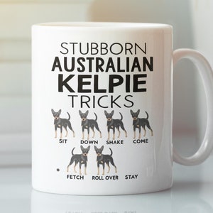 May include: White ceramic mug featuring the text "STUBBORN AUSTRALIAN KELPIE TRICKS" in black. Below the text are illustrations of an Australian Kelpie dog demonstrating tricks like sit, down, shake, and come. The mug has a curved handle.