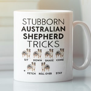 Peut inclure: Une tasse en céramique blanche avec un graphique noir et blanc d'un chien de berger australien. Le texte "Stubborn Australian Shepherd Tricks" est imprimé sur la tasse. Le chien est représenté en train d'effectuer diverses figures, notamment s'asseoir, se coucher, secouer, venir, chercher, rouler et rester.
