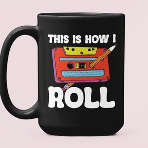 Może przedstawiać: Czarny kubek do kawy z białym nadrukiem kasety magnetofonowej z wystającym ołówkiem. Pod kasetą znajduje się napis "This is how I roll".