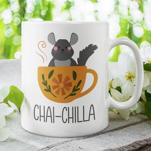 May include: White ceramic mug featuring a black and grey cartoon chinchilla sitting in a yellow teacup with a floral design. The text "CHAI-CHILLA" is printed on the mug.