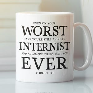 Könnte beinhalten: Weiße Keramik-Tasse mit schwarzem Text: "EVEN ON YOUR WORST DAYS YOU'RE STILL A GREAT INTERNIST AND AN AMAZING PERSON. DON'T YOU EVER FORGET IT!". Die Tasse hat einen gebogenen Henkel und steht auf einer reflektierenden Oberfläche.