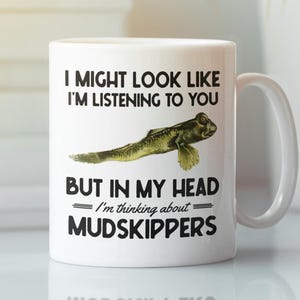 May include: White ceramic mug with a humorous design. The mug features the text "I MIGHT LOOK LIKE I'M LISTENING TO YOU BUT IN MY HEAD I'm thinking about MUDSKIPPERS" and a graphic of a mudskipper fish. The mug has a white handle.