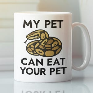 May include: White ceramic mug with the text "MY PET CAN EAT YOUR PET" in black. The mug features a brown and tan snake illustration. The mug has a white handle and is suitable for hot or cold beverages.