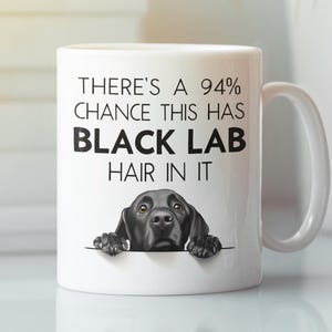 May include: White ceramic mug featuring a black Labrador illustration and the text "THERE'S A 94% CHANCE THIS HAS BLACK LAB HAIR IN IT". The mug has a curved handle. The background is blurred.