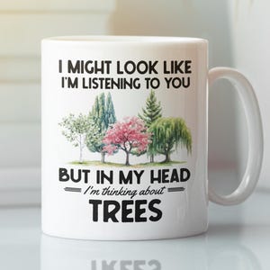 May include: White ceramic mug with a handle. The mug features the text "I MIGHT LOOK LIKE I'M LISTENING TO YOU BUT IN MY HEAD I'm thinking about TREES" along with a watercolor illustration of various trees.